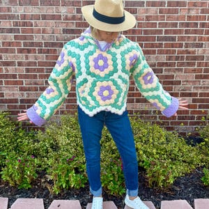May include: A hooded pullover featuring a quilted floral pattern in pastel yellow, light green, and lavender. The garment has long sleeves with lavender cuffs, a front kangaroo pocket, and a scalloped hem. It is paired with blue denim jeans and white sneakers.