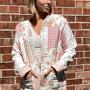 May include: A quilted jacket with a patchwork design. The jacket features a mix of floral and plaid patterns in shades of white, pink, and red. It has long sleeves, pockets, and a quilted texture. The jacket is worn open.