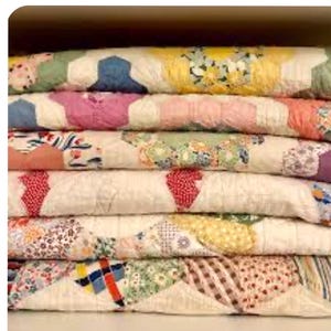 May include: A stack of handmade quilts with a variety of colorful, geometric patterns. The quilts feature a mix of floral, solid, and patterned fabrics in shades of pink, blue, yellow, and green. The quilts are neatly folded and stacked.