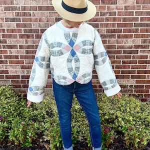 May include: A long-sleeved quilted pullover featuring a white base with a blue and pink interlocking ring pattern, paired with dark blue denim jeans, white sneakers, and a tan straw fedora hat with a black band.
