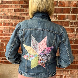 May include: A denim jacket with a colorful patchwork star design on the back. The star is made of various floral and patterned fabrics in shades of pink, blue, green, and yellow. The jacket is a classic style with button closures.