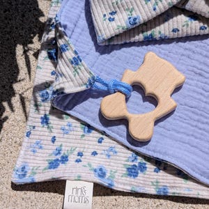 Könnte beinhalten: Ein Babydecken- und Mützen-Set mit blauem Blumenmuster auf einem hellgrauen Rippstoff. Eine hellblaue Musselindecke und ein Holzbeißring sind ebenfalls enthalten. Das Etikett lautet "riri & moms".