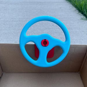 May include: A bright blue toy steering wheel with a red center and mounting bracket. The steering wheel is mounted on a cardboard box, suggesting a child's play vehicle. The design is simple and colorful, suitable for imaginative play.