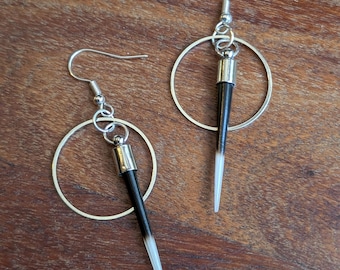 Pode incluir: Um par de brincos pendentes com ganchos de metal prateado, aros circulares finos e um espigão em forma de ponta. O espigão é preto no topo e termina numa ponta branca, fixado por uma tampa de metal prateado.