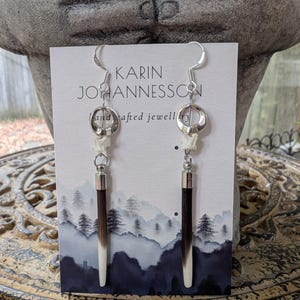 Könnte beinhalten: Hängeohrringe mit silberfarbenen Haken, kreisförmigen Metallringen, kleinen weißen Knochensegmenten und langen, spitzen Stachelschweinstacheln mit dunkelbraunen und weißen Spitzen. Präsentiert auf einer Karin Johannesson Karte.
