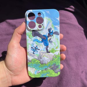 May include: A blue and white phone case featuring a cartoon illustration of a Lucario character in a fighting pose. The case has a landscape background with rocks, green foliage, and a blue sky.