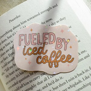 Può includere: Un adesivo con la scritta "FUELED BY iced coffee" in un font giocoso. L'adesivo ha uno sfondo rosa chiaro con accenti olografici e una forma ondulata, posto su un libro aperto.