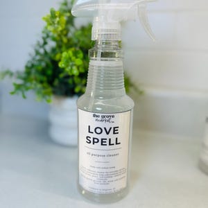 May include: Clear spray bottle with a white label that reads "LOVE SPELL all purpose cleaner" in black text. The bottle has a clear spray nozzle and is partially filled with a clear liquid. A green plant is in the background.