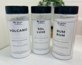 May include: Three white plastic containers with black lids, labeled "VOLCANIC," "SOL LUXE," and "BUM BUM." Each container has a white label with black text, advertising "upholstery deodorizer." The background is a blurred, light-colored surface.