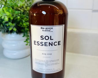 May include: A brown bottle of "SOL ESSENCE" mop soap with a black cap. The label includes the text "the grove market co." and instructions. A green plant in a white pot is in the background.