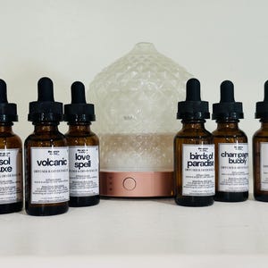 Puede incluir: Un difusor de aceites esenciales con una base de oro rosa y una cúpula transparente texturizada, flanqueado por seis botellas de vidrio ámbar con cuentagotas negros. Las botellas están etiquetadas con nombres como "Sol Luxe" y "Winter Apple".