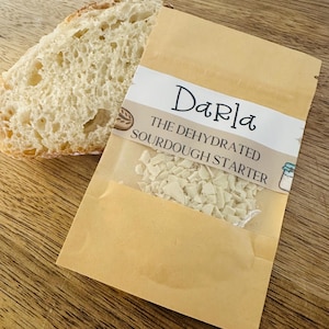 Może przedstawiać: Kawałek chleba obok opakowania odwodnionego zakwasu. Na beżowym opakowaniu widnieje napis "Darla" oraz "THE DEHYDRATED SOURDOUGH STARTER". Zakwas jest widoczny przez przezroczyste okienko.
