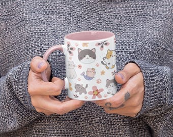 Può includere: Tazza in ceramica bianca con interno e manico rosa, ornata da un motivo ripetuto di gatti dei cartoni animati, fiori e impronte di zampe. I gatti sono in varie pose e colori, creando un design stravagante. La tazza è tenuta da una persona.