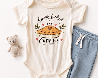 May include: A cream-colored short-sleeve bodysuit featuring a graphic of a baked pie with a heart on top, surrounded by small red hearts and green leaves, with the text "home baked CUTIE PIE." A small crocheted teddy bear and blue pants.