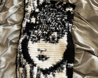 May include: A handmade crochet piece featuring a face design. The artwork uses black, white, and gray yarn to create the image of a face. The piece is rectangular and appears to be a decorative item or a small textile.