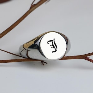 May include: A polished silver signet ring with a white face and a black letter "J" in a gothic font. The ring is oval-shaped and rests on a brown twig against a white background. The ring is a piece of jewellery.