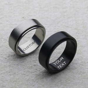 Può includere: Due anelli, uno argentato e uno nero, sono esposti su una superficie strutturata. L'anello argentato ha la scritta "CUSTOM" incisa all'interno, mentre l'anello nero ha "YOUR TEXT". Entrambi gli anelli hanno un design a fascia piatta e larga.