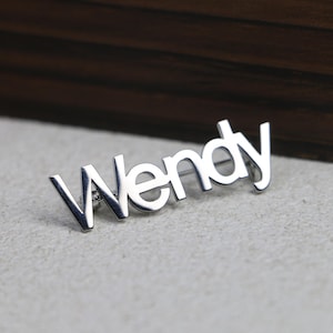 Puede incluir: Un pin plateado con el nombre "Wendy" en una fuente estilizada. El pin está sobre una superficie gris claro, con un objeto de madera marrón oscuro en el fondo. Probablemente sea un accesorio personalizado.