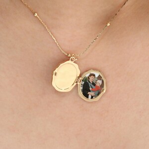 Pode incluir: Um colar com um pingente de medalh&atilde;o oval dourado, aberto para revelar uma pequena fotografia. O medalh&atilde;o pende de uma delicada corrente com pequenas contas redondas. A fotografia mostra uma pessoa segurando um beb&ecirc;.