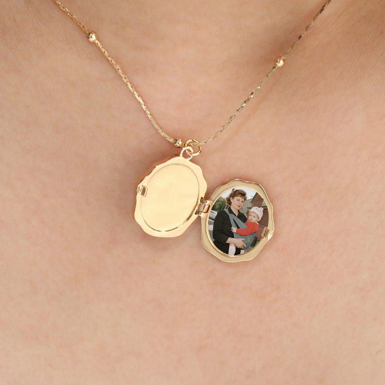 Pode incluir: Um colar com um pingente de medalh&atilde;o oval dourado, aberto para revelar uma pequena fotografia. O medalh&atilde;o pende de uma delicada corrente com pequenas contas redondas. A fotografia mostra uma pessoa segurando um beb&ecirc;.