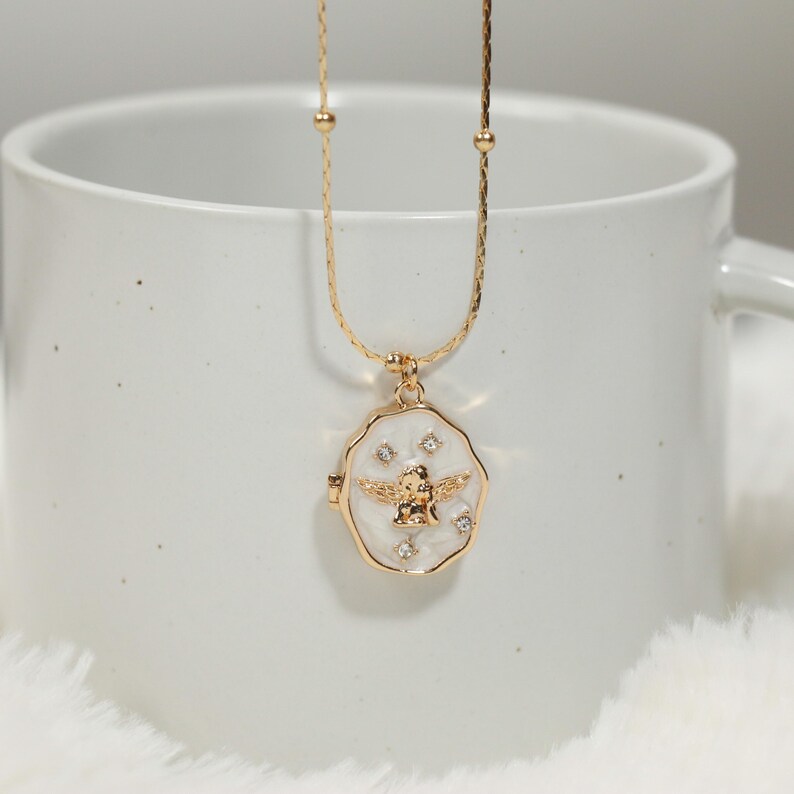 Pode incluir: Colar dourado com um pingente com fundo de esmalte branco e um design de anjo dourado. O pingente &eacute; adornado com pequenos cristais brilhantes. O colar &eacute; exibido em uma caneca branca.