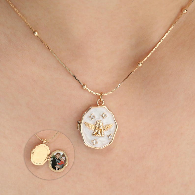 Pode incluir: Colar com pingente de medalh&atilde;o dourado com esmalte branco. O pingente apresenta um design de anjo dourado e pequenos detalhes de diamantes. Um medalh&atilde;o menor e aberto com uma foto &eacute; mostrado abaixo. A corrente &eacute; delicada.