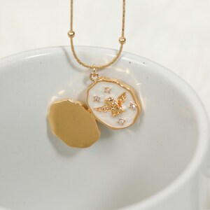 Pode incluir: Um colar dourado com um pingente. O pingente tem um fundo de esmalte branco e um design de anjo dourado, adornado com pequenos cristais. O colar &eacute; exibido em uma superf&iacute;cie branca.