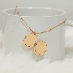 Pode incluir: Um colar com um medalh&atilde;o dourado em forma oval, aberto para revelar dois compartimentos. A corrente do colar &eacute; fina, adornada com pequenas contas redondas. O medalh&atilde;o est&aacute; dentro de uma caneca branca.
