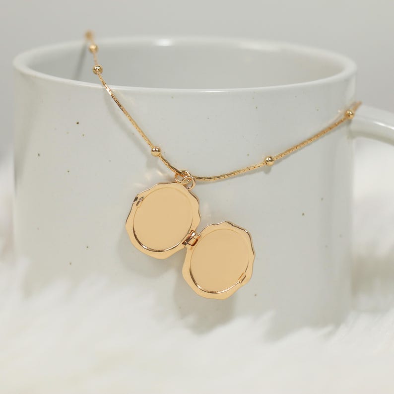 Pode incluir: Um colar com um medalh&atilde;o dourado em forma oval, aberto para revelar dois compartimentos. A corrente do colar &eacute; fina, adornada com pequenas contas redondas. O medalh&atilde;o est&aacute; dentro de uma caneca branca.