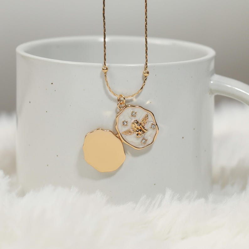 Pode incluir: Colar com pingente de medalh&atilde;o dourado com um anjo detalhado. O pingente tem um fundo de esmalte branco com um anjo dourado e pequenos cristais transparentes. O medalh&atilde;o est&aacute; aberto, revelando um interior dourado. O colar &eacute; exibido em uma caneca branca.