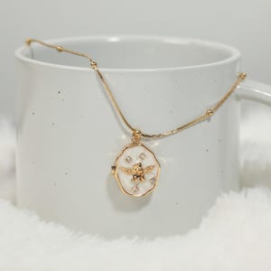 Pode incluir: Colar dourado com um pingente com um design de abelha. O pingente tem um fundo branco com uma abelha dourada e pequenos detalhes de cristal. O colar &eacute; exibido contra uma caneca branca e uma superf&iacute;cie macia e branca.