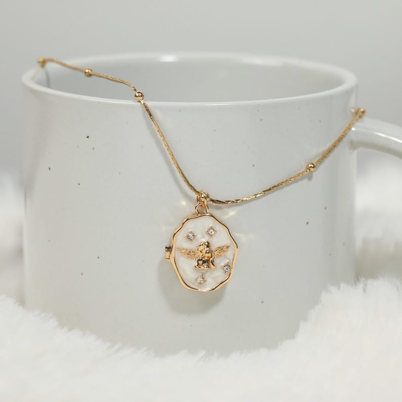 Pode incluir: Colar dourado com um pingente com um design de abelha. O pingente tem um fundo branco com uma abelha dourada e pequenos detalhes de cristal. O colar &eacute; exibido contra uma caneca branca e uma superf&iacute;cie macia e branca.
