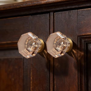 Puede incluir: Dos pomos de armario de cristal transparente y facetado con un suave tono rosado, montados sobre bases de latón. Los pomos son octogonales y están fijados a un armario de madera marrón oscuro. La ferretería añade un toque de elegancia al mueble.