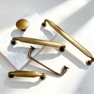 May include: An assortment of antique brass cabinet hardware, including a round knob, two long pulls, a shorter pull, and a curved handle. The hardware is arranged on a white surface, showcasing the aged finish and various designs.