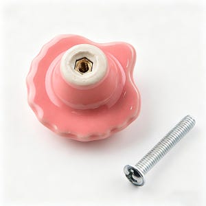 May include: A pink ceramic cabinet knob with a scalloped edge and a white base. A silver screw is positioned next to the knob. The knob has a glossy finish and a decorative design, suitable for furniture.