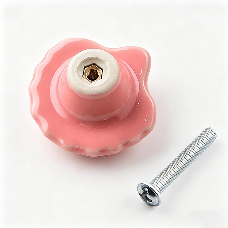 May include: A pink ceramic cabinet knob with a scalloped edge and a white base. A silver screw is positioned next to the knob. The knob has a glossy finish and a decorative design, suitable for furniture.