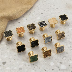 K&ouml;nnte beinhalten: Eine Sammlung dekorativer Schrankkn&ouml;pfe mit einem vierbl&auml;ttrigen Kleeblatt-Design. Die Kn&ouml;pfe weisen verschiedene Farben und Texturen auf, darunter Schwarz, Wei&szlig;, Gold und Gr&uuml;n. Jeder Knopf hat einen goldfarbenen Sockel.