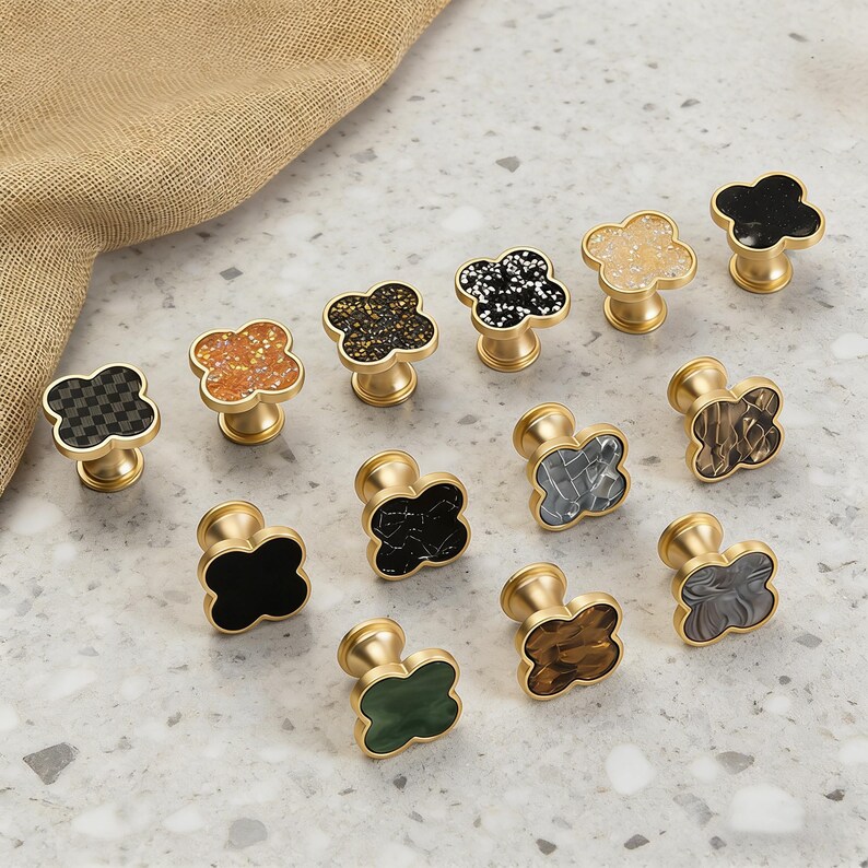 K&ouml;nnte beinhalten: Eine Sammlung dekorativer Schrankkn&ouml;pfe mit einem vierbl&auml;ttrigen Kleeblatt-Design. Die Kn&ouml;pfe weisen verschiedene Farben und Texturen auf, darunter Schwarz, Wei&szlig;, Gold und Gr&uuml;n. Jeder Knopf hat einen goldfarbenen Sockel.