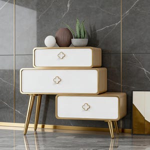 K&ouml;nnte beinhalten: Ein dreistufiger wei&szlig;-goldener Beistelltisch mit kleeblattf&ouml;rmigen Schubladengriffen. Der Tisch hat goldene Beine und Zierleisten. Dekorative Gegenst&auml;nde, darunter eine Vase und eine Pflanze, stehen oben.