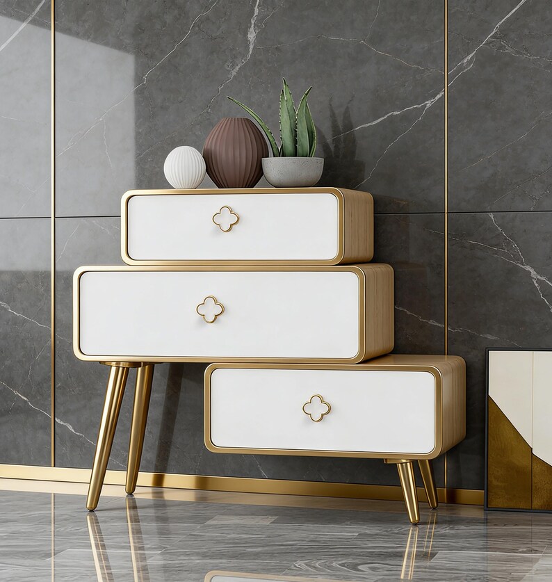 K&ouml;nnte beinhalten: Ein dreistufiger wei&szlig;-goldener Beistelltisch mit kleeblattf&ouml;rmigen Schubladengriffen. Der Tisch hat goldene Beine und Zierleisten. Dekorative Gegenst&auml;nde, darunter eine Vase und eine Pflanze, stehen oben.