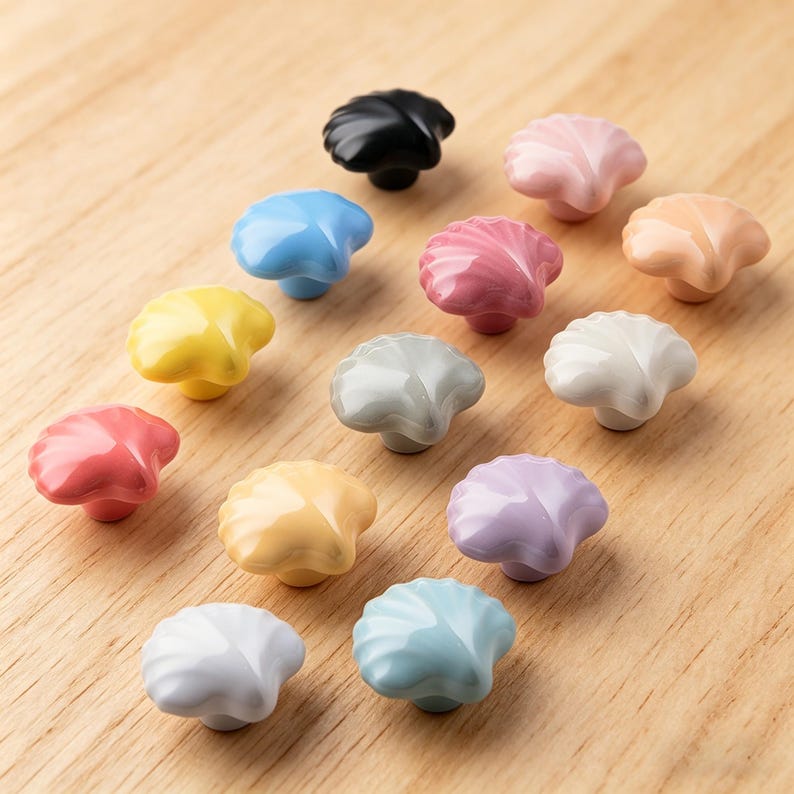 May include: A selection of seashell-shaped cabinet knobs in a range of colours, including black, pink, blue, yellow, and white. The knobs are displayed on a light brown wooden surface. Each knob has a unique colour and glossy finish.