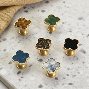 May include: Six decorative cabinet knobs with a cloverleaf design. The knobs have a gold-coloured base and feature various colours and patterns, including brown, green, black, blue, and grey. The knobs are arranged on a light grey surface.