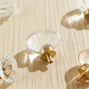May include: Clear glass oval cabinet knobs with brass bases are arranged on a light wood surface. The knobs have a smooth, rounded shape, and the brass bases add a touch of elegance. The knobs are designed for furniture.