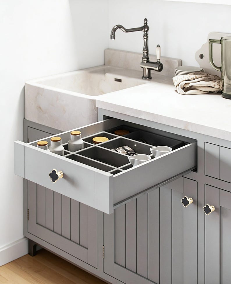K&ouml;nnte beinhalten: Ein grauer K&uuml;chenschrank mit einer offenen Schublade, die eine organisierte Aufbewahrung zeigt. Die Schublade enth&auml;lt kleine Gl&auml;ser, Utensilien und Schalen. Der Schrank hat schwarze und goldfarbene Kleeblattkn&ouml;pfe. Ein wei&szlig;es Waschbecken und eine Armatur sind im Hintergrund zu sehen.