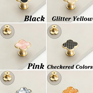 K&ouml;nnte beinhalten: Eine Auswahl an dekorativen Schrankkn&ouml;pfen in Kleeblattform mit goldenen Basen. Die Kn&ouml;pfe sind in verschiedenen Farben erh&auml;ltlich, darunter Schwarz, Glitzergelb, Pink, Schachbrettmuster, Auroragrau und Glitzerorange. Text kennzeichnet jede Farbe.