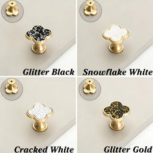 K&ouml;nnte beinhalten: Vier Schrankkn&ouml;pfe mit Kleeblatt-Design. Die Kn&ouml;pfe sind in vier verschiedenen Ausf&uuml;hrungen erh&auml;ltlich: Glitter Black, Snowflake White, Cracked White und Glitter Gold. Jeder Knopf hat einen goldfarbenen Sockel und eine dekorative Oberseite.