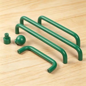 Puede incluir: Un conjunto de herrajes verdes para armarios, que incluye tres tiradores de barra, un pomo redondo y un pequeño pomo cilíndrico. Los herrajes están dispuestos sobre una superficie de madera clara, mostrando un diseño moderno.