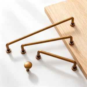Puede incluir: Un conjunto de cuatro piezas de herrajes de latón antiguo para armarios. El conjunto incluye tres tiradores de barra de diferentes longitudes y un pomo redondo. El hardware se muestra sobre una superficie blanca con una tabla de madera en el fondo.