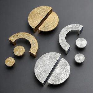 May include: An assortment of gold and silver-toned cabinet hardware. The collection includes half-circle handles, circular knobs, and split-circle handles, all with a textured, sunburst-like pattern. The hardware is displayed on a dark gray surface.