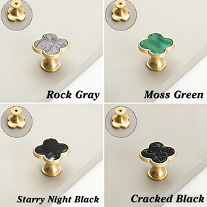 K&ouml;nnte beinhalten: Vier Schrankkn&ouml;pfe in Rock Gray, Moss Green, Starry Night Black und Cracked Black. Jeder Knopf hat eine kleeblattf&ouml;rmige Oberseite mit dekorativem Finish und eine goldfarbene Basis. Der Text darunter zeigt die Farbe an.